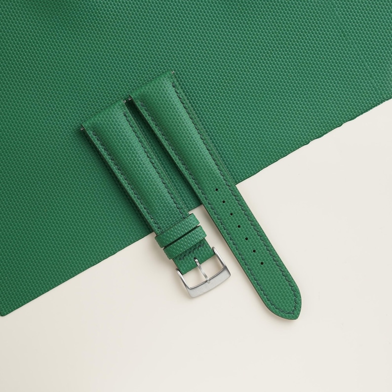 May include: A pair of green watch straps with a textured surface. The straps are made of a durable material and feature a silver-colored buckle. The straps are displayed on a white and green background.