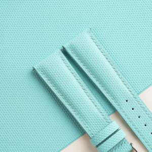 Puede incluir: Primer plano de una correa de reloj azul claro con una superficie texturizada. La correa tiene un borde cosido y una hebilla plateada. El fondo es una superficie texturizada azul claro a juego.