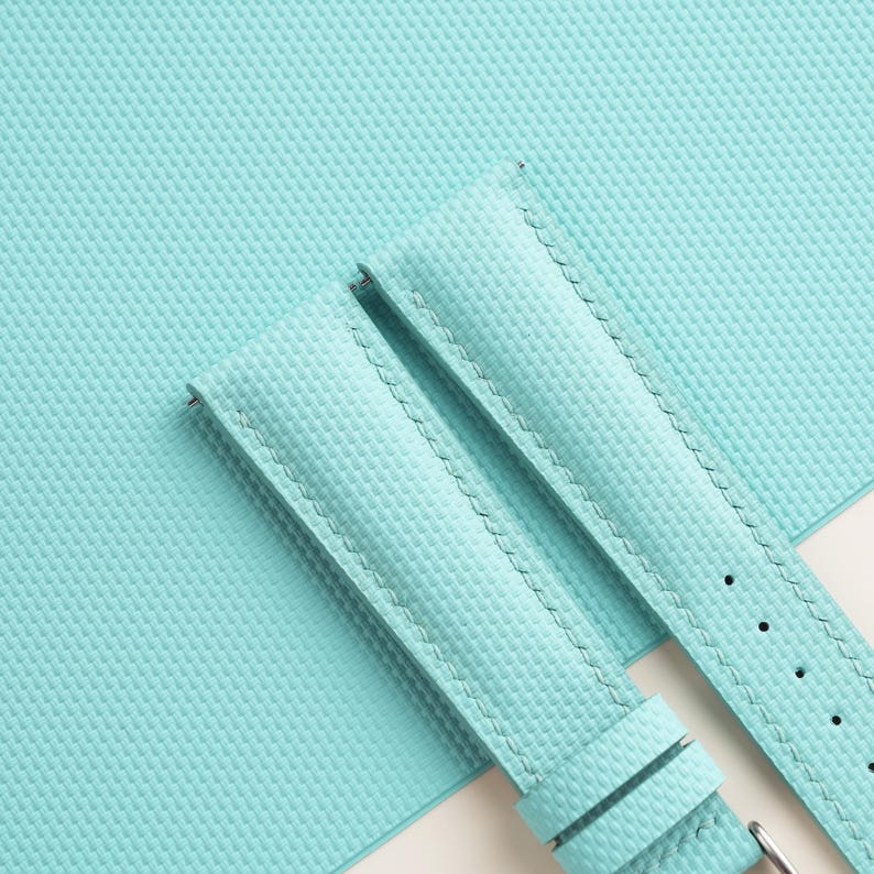 Puede incluir: Primer plano de una correa de reloj azul claro con una superficie texturizada. La correa tiene un borde cosido y una hebilla plateada. El fondo es una superficie texturizada azul claro a juego.