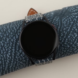 May include: A black smartwatch with a round face and a white and blue patterned band. The watch is on a blue patterned surface.