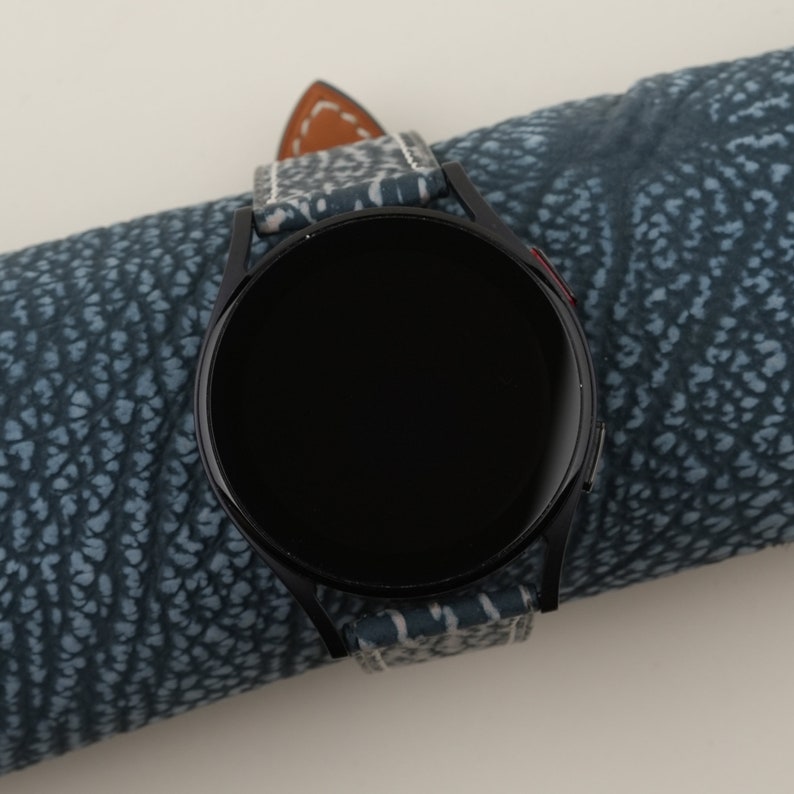 May include: A black smartwatch with a round face and a white and blue patterned band. The watch is on a blue patterned surface.