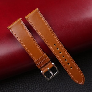 May include: Two brown leather watch straps with white stitching. The straps are laid out on a red surface. One strap has a silver buckle.