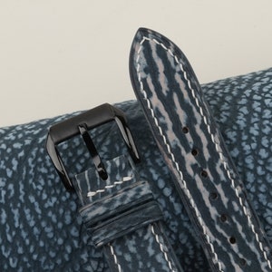 May include: A blue leather watch strap with white stitching and a black buckle. The strap has a textured, patterned surface.