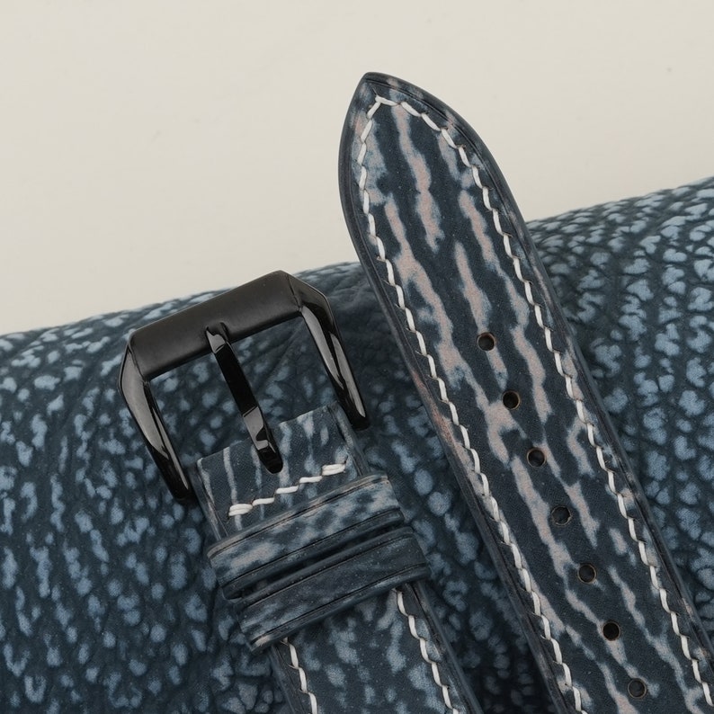 May include: A blue leather watch strap with white stitching and a black buckle. The strap has a textured, patterned surface.