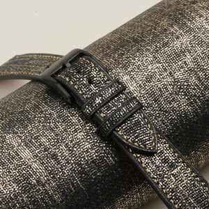 K&ouml;nnte beinhalten: Ein schwarzes Lederarmband mit goldenen N&auml;hten. Das Armband ist um eine gold-schwarze gemusterte Oberfl&auml;che gewickelt.