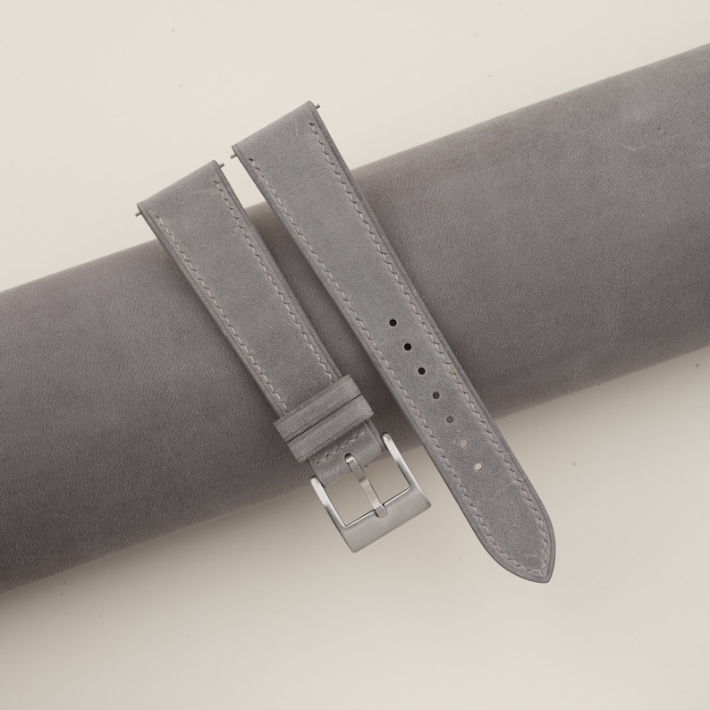 May include: Two gray leather watch straps with a silver buckle. The straps are laid out on a gray fabric surface.