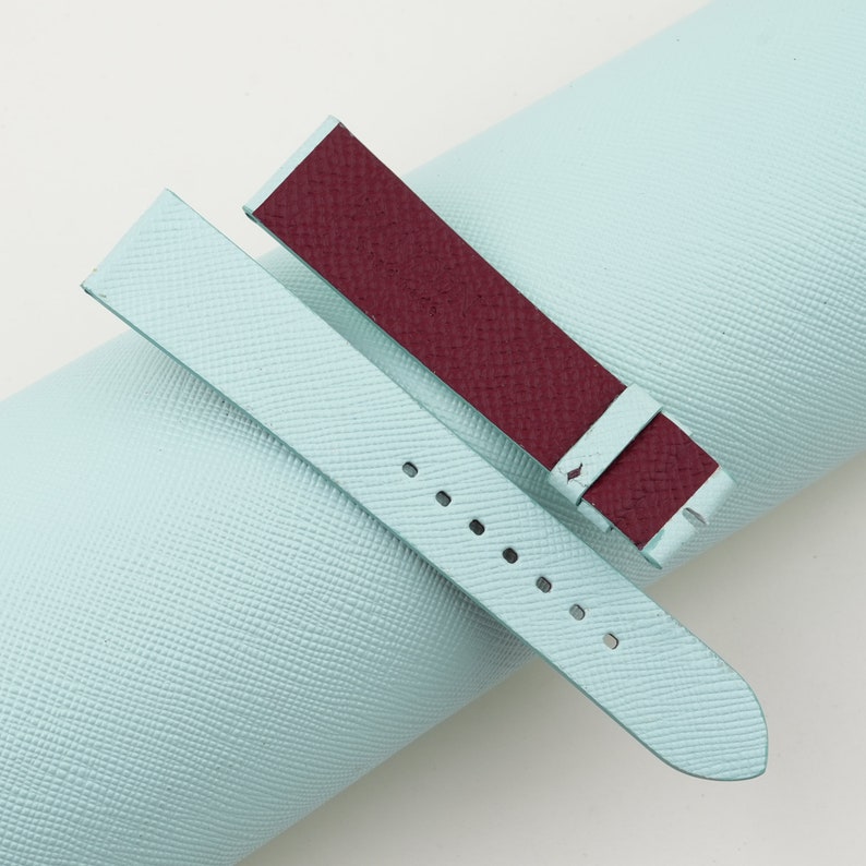 May include: Two watch straps, one light blue and one burgundy, are laid out on a light blue textured surface. The straps are made of leather and have a simple design.