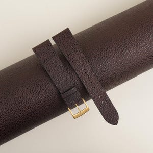 May include: A brown leather watch strap with a gold buckle. The strap is rolled up and the buckle is visible.