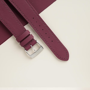 May include: A burgundy watch strap with a textured surface and a silver-colored buckle. The strap is made of a durable material and features stitching along the edges. The strap is designed to be attached to a watch.
