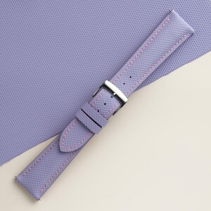 Puede incluir: Una correa de reloj lavanda con costuras rosas y una hebilla plateada. La correa tiene una superficie texturizada y est&aacute; sobre un fondo lavanda.