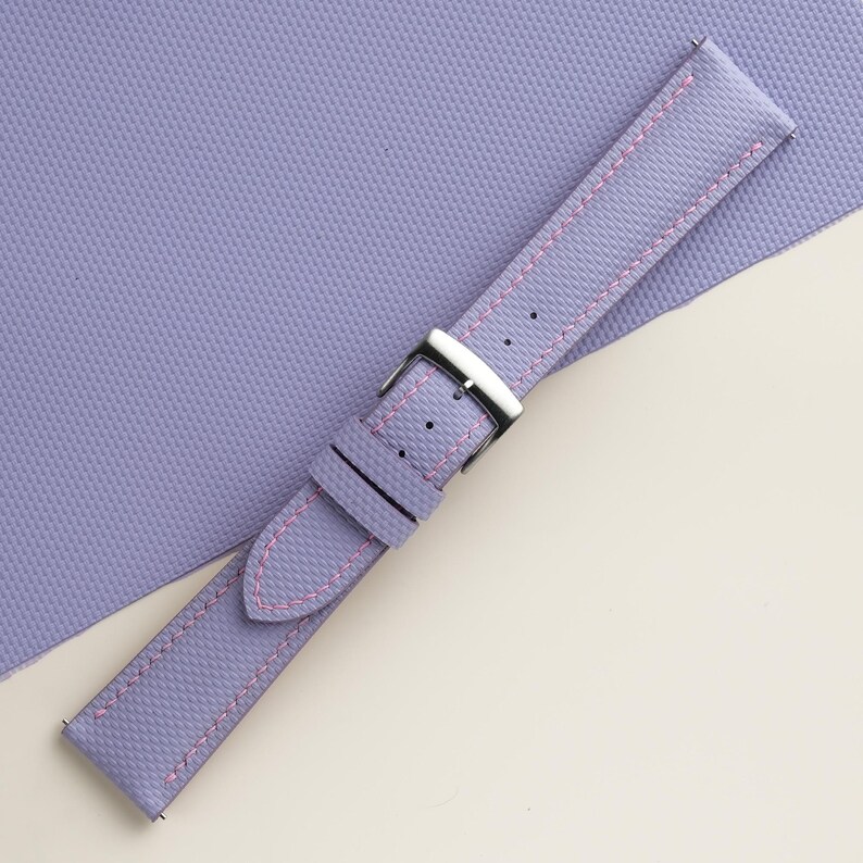 Puede incluir: Una correa de reloj lavanda con costuras rosas y una hebilla plateada. La correa tiene una superficie texturizada y est&aacute; sobre un fondo lavanda.