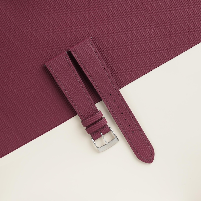May include: Burgundy watch strap with a textured surface and a silver-colored buckle. The strap is made of a durable material and is designed to be attached to a watch. The background is a combination of burgundy and white.