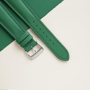 May include: Close-up of a green watch strap with a textured surface and a silver buckle. The strap is stitched with dark green thread and lies on a white and green background. The strap has several holes for adjustment.