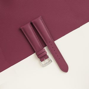 May include: A pair of burgundy watch straps with a textured surface and a silver-colored buckle. The straps are displayed on a burgundy textured background and a white surface.