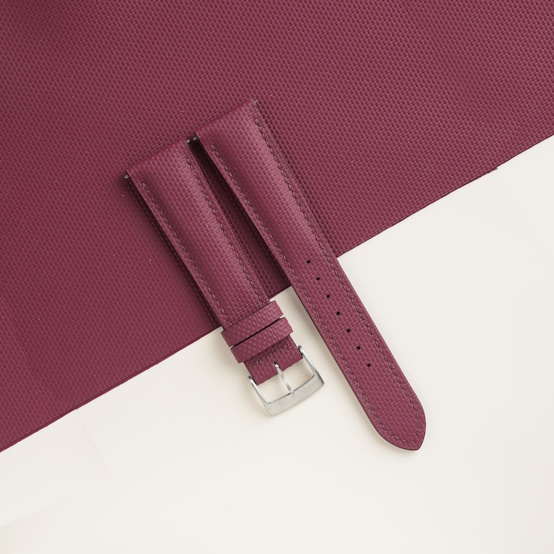 May include: A pair of burgundy watch straps with a textured surface and a silver-colored buckle. The straps are displayed on a burgundy textured background and a white surface.