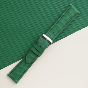 May include: A green watch strap with a textured surface and a silver-colored buckle. The strap is made of a durable material and features stitching along the edges. The watch band is angled across a green surface.