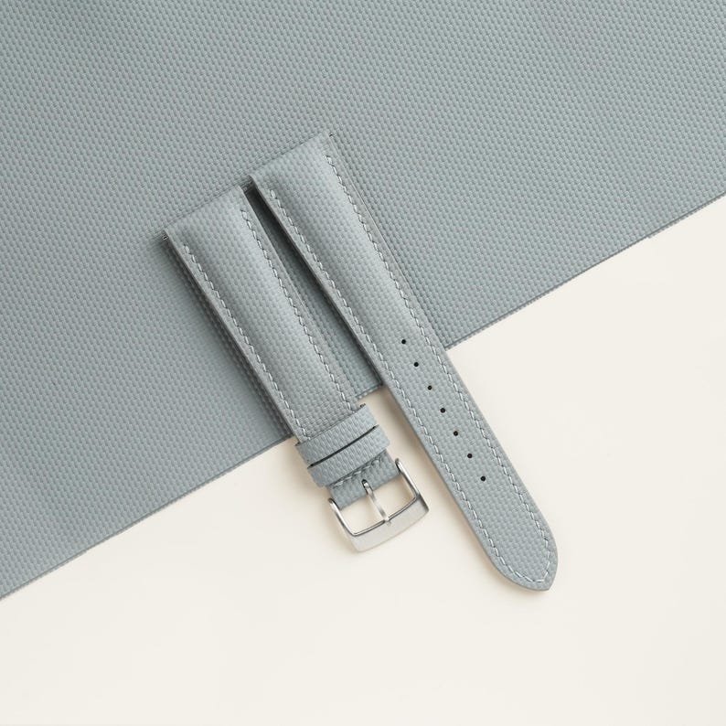 Puede incluir: Una correa de reloj gris claro con una superficie texturizada y una hebilla plateada. La correa est&aacute; hecha de un material duradero y presenta costuras en los bordes. La correa del reloj se muestra sobre un fondo gris claro y blanco.