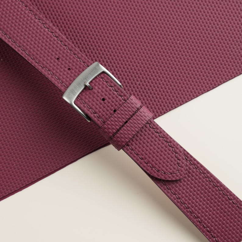 May include: Close-up of a burgundy watch strap with a textured surface and a silver-colored buckle. The strap is stitched with matching thread and lies on a white and burgundy background.