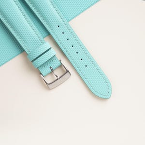 Puede incluir: Primer plano de una correa de reloj azul claro con una superficie texturizada y una hebilla plateada. La correa tiene un borde cosido y varios agujeros para ajustar. La correa del reloj es de color turquesa claro.