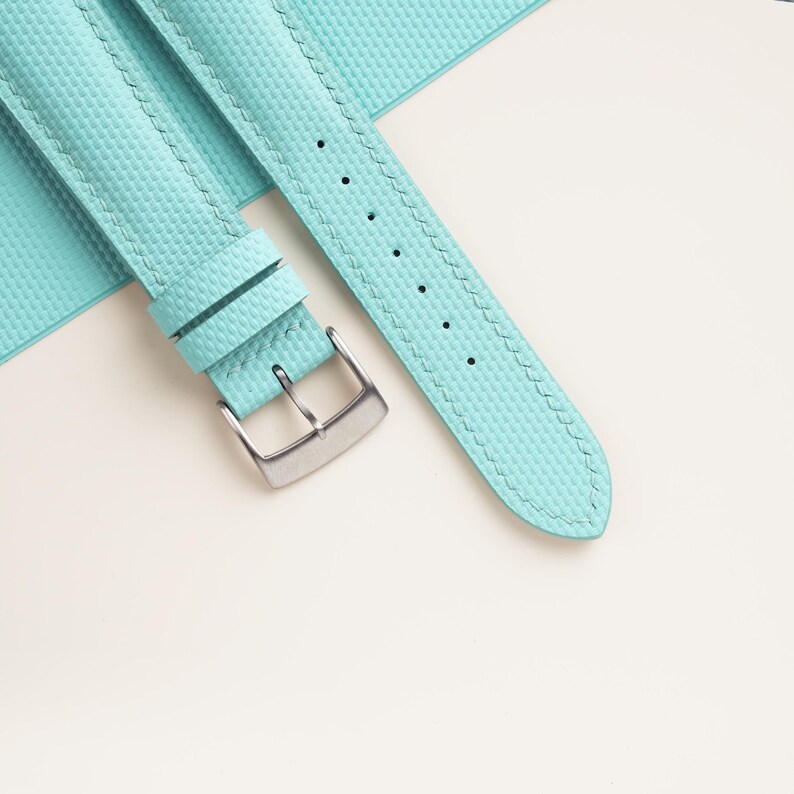 Puede incluir: Primer plano de una correa de reloj azul claro con una superficie texturizada y una hebilla plateada. La correa tiene un borde cosido y varios agujeros para ajustar. La correa del reloj es de color turquesa claro.