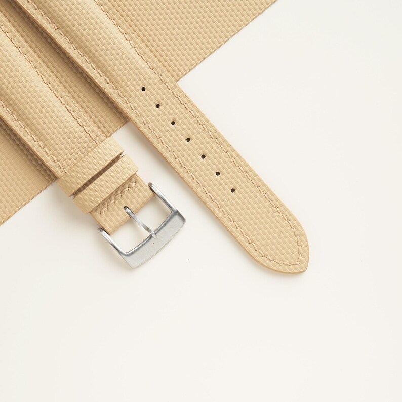 Puede incluir: Correa de reloj beige con superficie texturizada y hebilla plateada. La correa est&aacute; hecha de un material duradero y presenta costuras en los bordes. La correa del reloj se muestra sobre un fondo blanco.