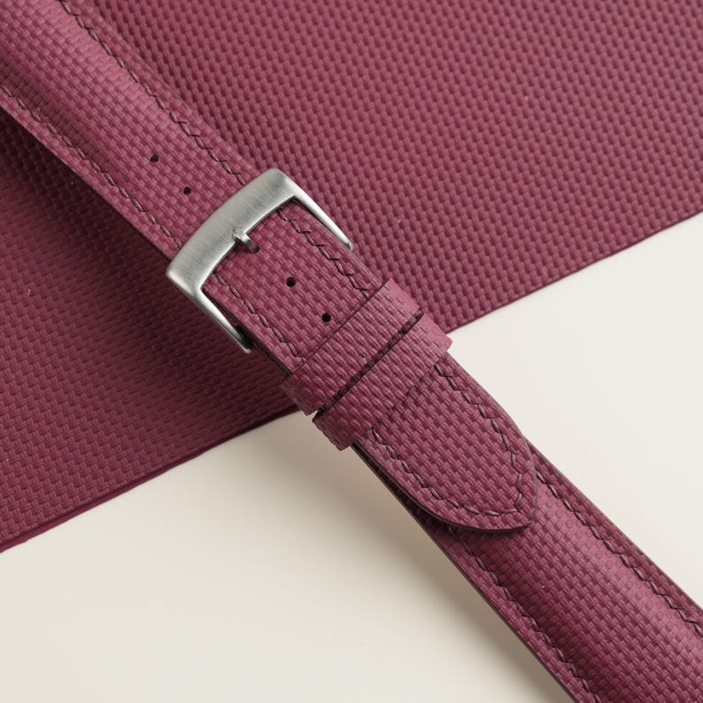 May include: Close-up of a burgundy watch strap with a textured surface and a silver-colored buckle. The strap is stitched with matching thread and lies against a white background.