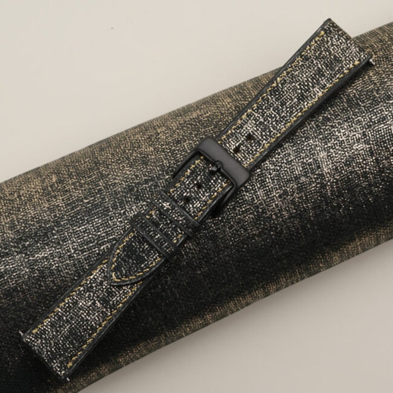 K&ouml;nnte beinhalten: Ein schwarzes Lederarmband mit goldenen N&auml;hten. Das Armband liegt auf einer gold-schwarzen, strukturierten Oberfl&auml;che.