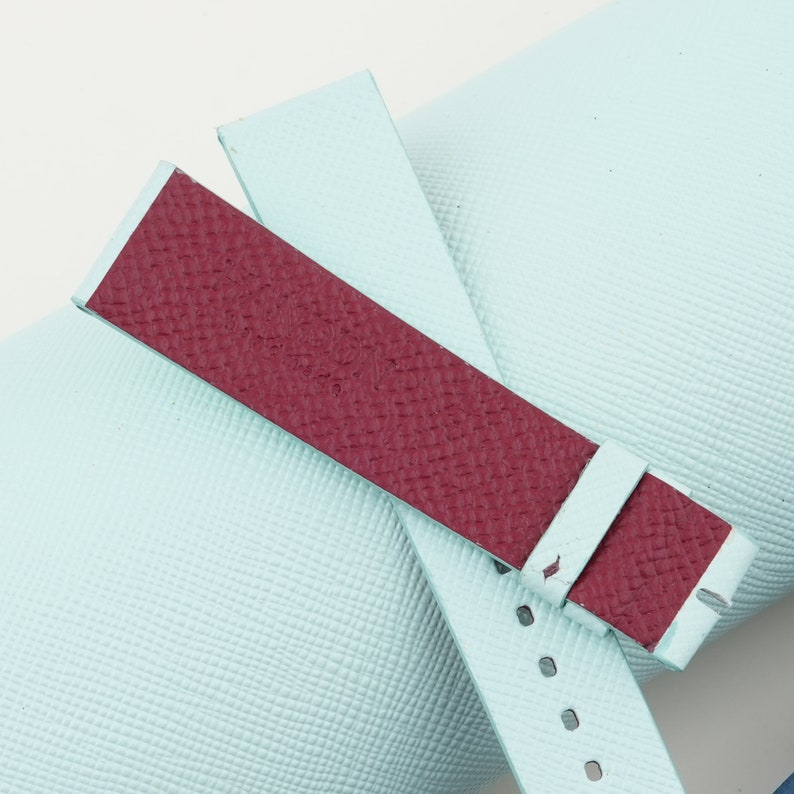 May include: A light blue and burgundy leather watch strap with a textured finish.