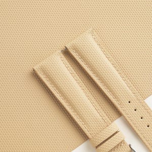 Puede incluir: Correa de reloj de color beige con superficie texturizada y costuras en contraste. La correa tiene una hebilla y tres orificios de ajuste. El fondo es de color beige a juego.