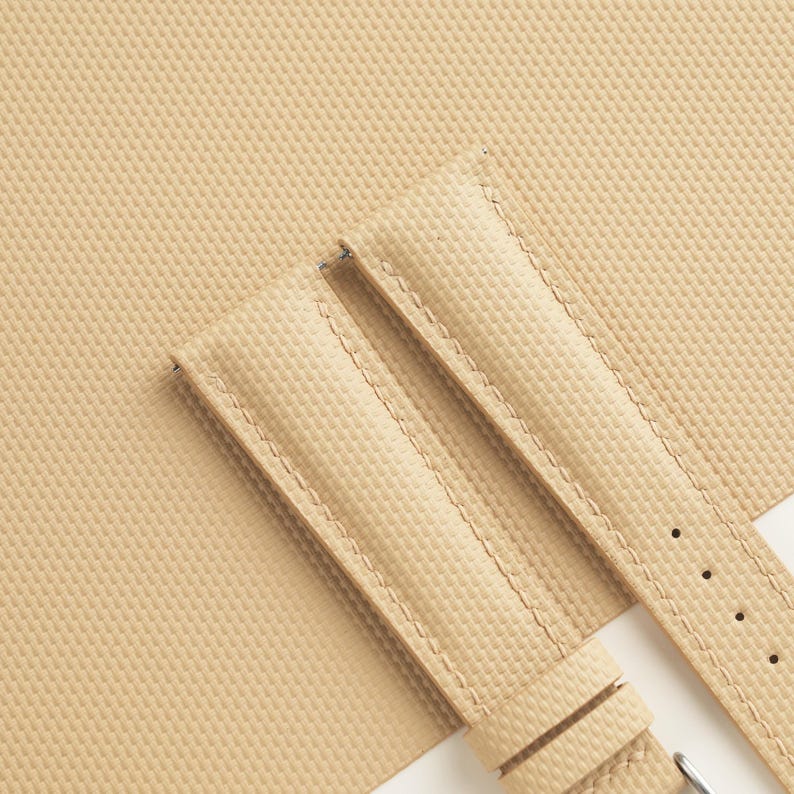 Puede incluir: Correa de reloj de color beige con superficie texturizada y costuras en contraste. La correa tiene una hebilla y tres orificios de ajuste. El fondo es de color beige a juego.