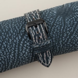 May include: A blue and white leather watch strap with a black buckle. The strap is rolled up on a blue patterned surface.