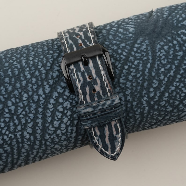 May include: A blue and white leather watch strap with a black buckle. The strap is rolled up on a blue patterned surface.