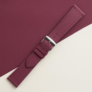 May include: A burgundy watch strap with a textured surface and a silver-colored buckle. The strap is angled across a matching burgundy surface, creating a visually appealing contrast. The watch strap is a stylish accessory.