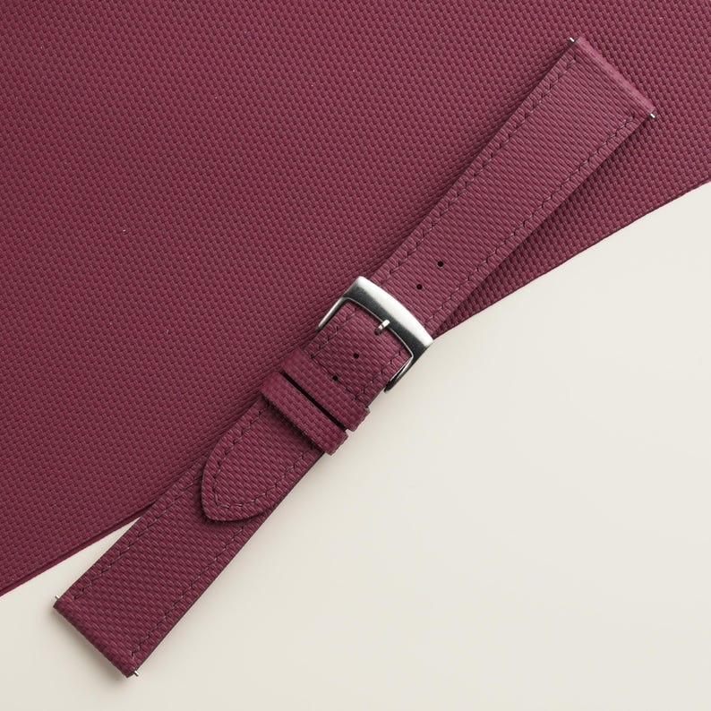 May include: A burgundy watch strap with a textured surface and a silver-colored buckle. The strap is angled across a matching burgundy surface, creating a visually appealing contrast. The watch strap is a stylish accessory.