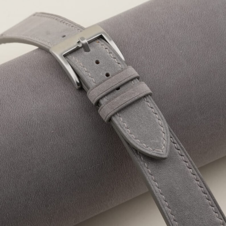 May include: A gray suede watch strap with a silver buckle. The strap has a stitched edge and a textured surface.