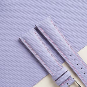 Puede incluir: Dos correas de reloj lavanda con costuras rosas se muestran sobre un fondo texturizado a juego. Las correas tienen un dise&ntilde;o cl&aacute;sico con una hebilla y orificios de ajuste. La est&eacute;tica general es limpia y moderna, adecuada para varios estilos de relojes.