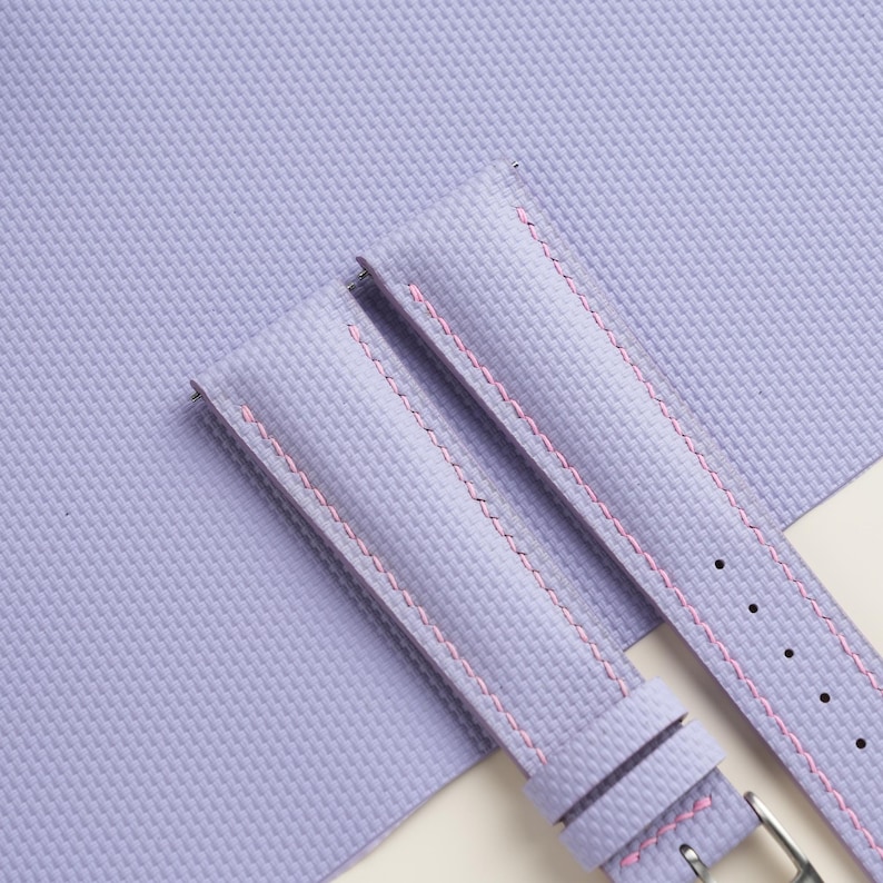 Puede incluir: Dos correas de reloj lavanda con costuras rosas se muestran sobre un fondo texturizado a juego. Las correas tienen un dise&ntilde;o cl&aacute;sico con una hebilla y orificios de ajuste. La est&eacute;tica general es limpia y moderna, adecuada para varios estilos de relojes.