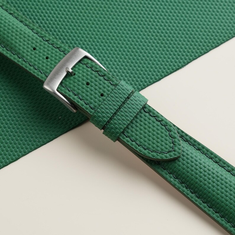 May include: Close-up of a green watch strap with a textured weave pattern. The strap features a silver-colored buckle and matching stitching. The strap is displayed on a white and green background.
