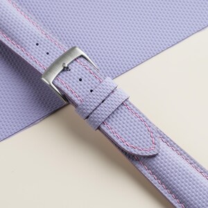 Puede incluir: Primer plano de una correa de reloj lavanda con costuras rosas y una hebilla plateada. La correa tiene un patr&oacute;n de tejido texturizado. El fondo es de material lavanda a juego.
