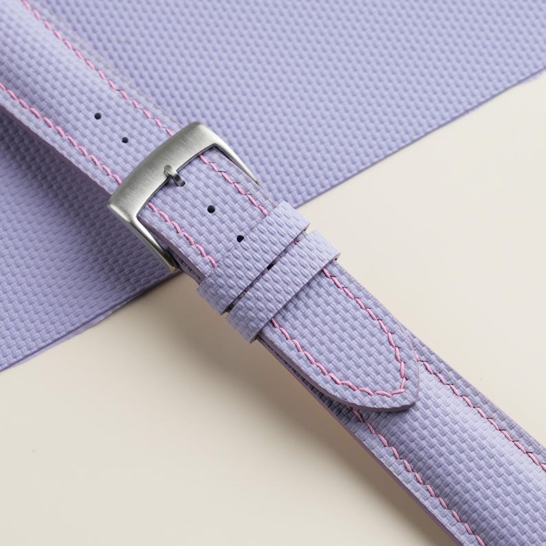 Puede incluir: Primer plano de una correa de reloj lavanda con costuras rosas y una hebilla plateada. La correa tiene un patr&oacute;n de tejido texturizado. El fondo es de material lavanda a juego.