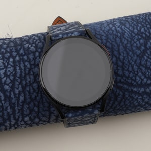 May include: A black smartwatch with a round face and a blue leather strap. The watch is lying on a blue textured surface.