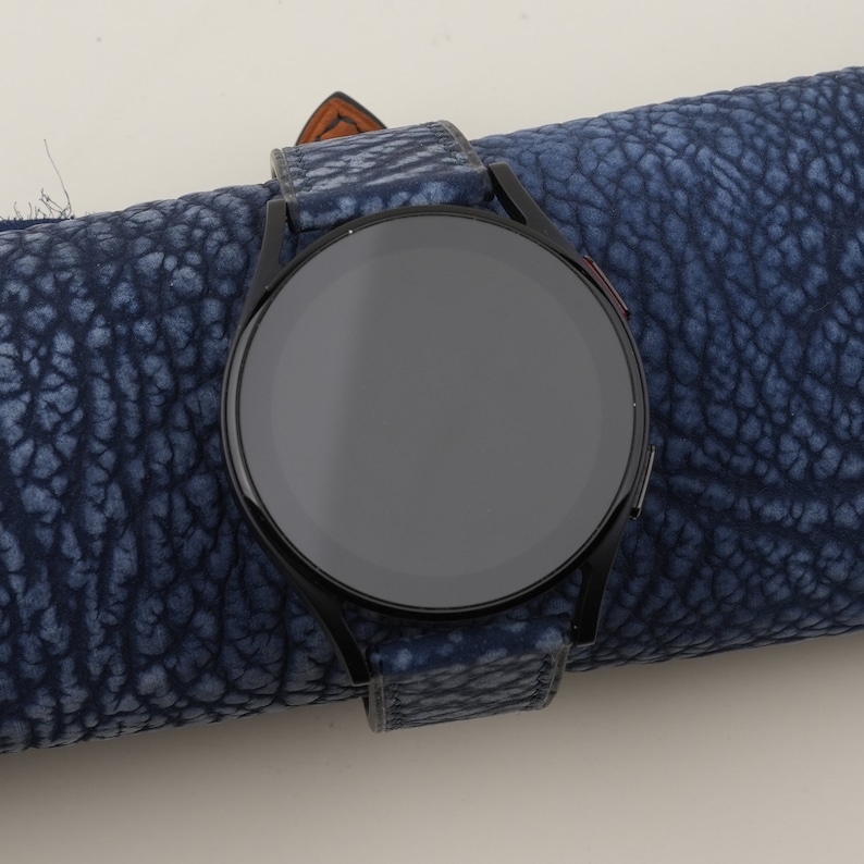 May include: A black smartwatch with a round face and a blue leather strap. The watch is lying on a blue textured surface.
