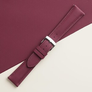 May include: A burgundy watch strap with a textured pattern and a silver-colored buckle. The strap is made of a durable material and is designed to fit a watch. The background is a matching burgundy color.