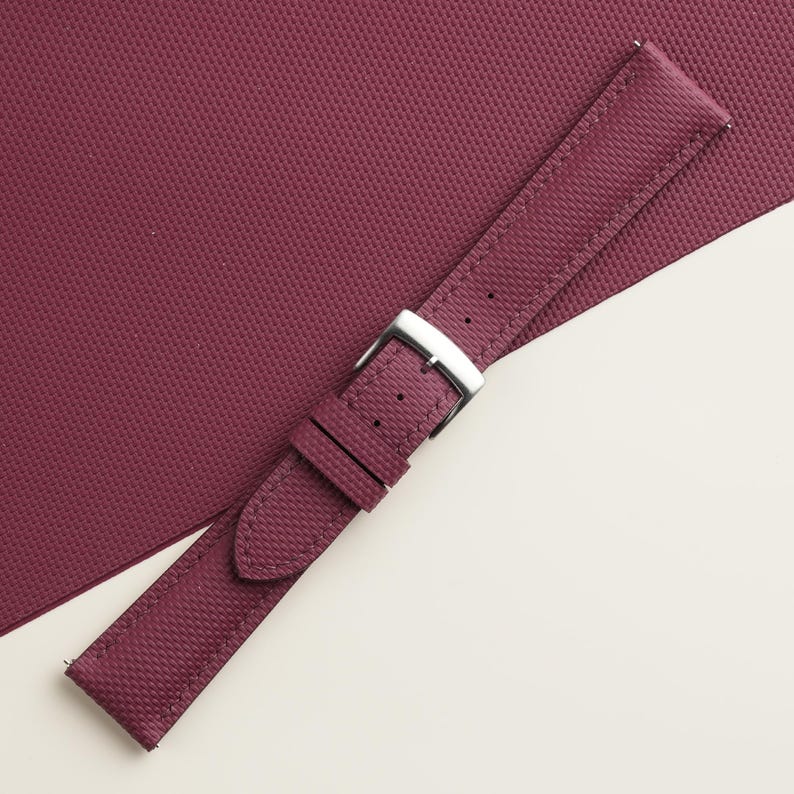 May include: A burgundy watch strap with a textured pattern and a silver-colored buckle. The strap is made of a durable material and is designed to fit a watch. The background is a matching burgundy color.