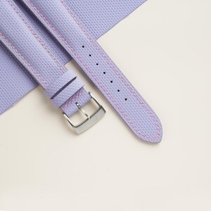 Puede incluir: Primer plano de una correa de reloj lavanda con costuras rosas. La correa tiene una superficie texturizada y una hebilla plateada. La correa del reloj se muestra sobre un fondo blanco.