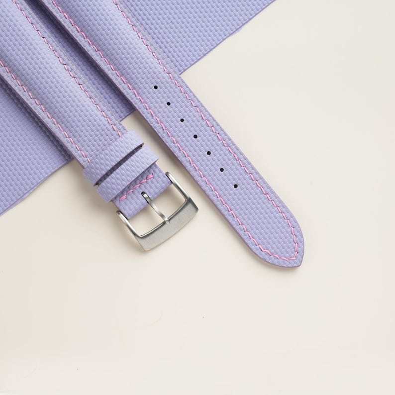 Puede incluir: Primer plano de una correa de reloj lavanda con costuras rosas. La correa tiene una superficie texturizada y una hebilla plateada. La correa del reloj se muestra sobre un fondo blanco.