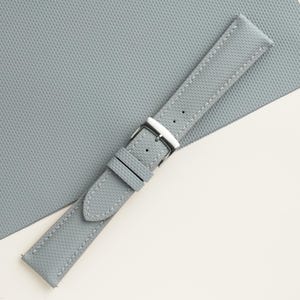 Puede incluir: Una correa de reloj gris claro con costuras blancas y una hebilla plateada. La correa tiene una superficie texturizada y est&aacute; en &aacute;ngulo sobre un fondo gris claro.