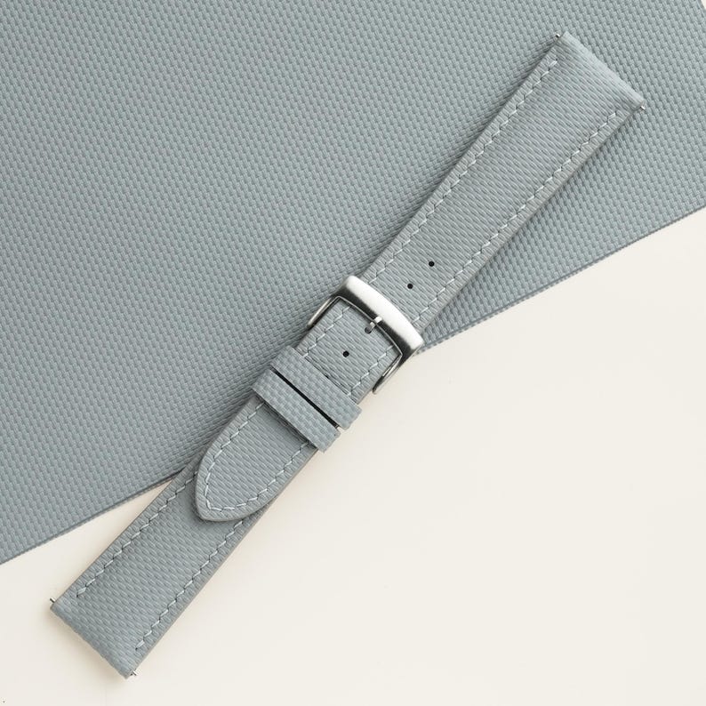 Puede incluir: Una correa de reloj gris claro con costuras blancas y una hebilla plateada. La correa tiene una superficie texturizada y est&aacute; en &aacute;ngulo sobre un fondo gris claro.