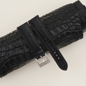 May include: Black alligator leather watch strap with a silver buckle. The strap is rolled up and the buckle is visible.
