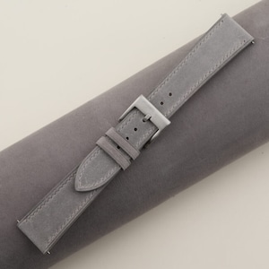 May include: A gray suede watch strap with a silver buckle. The strap is rolled up on a gray surface.